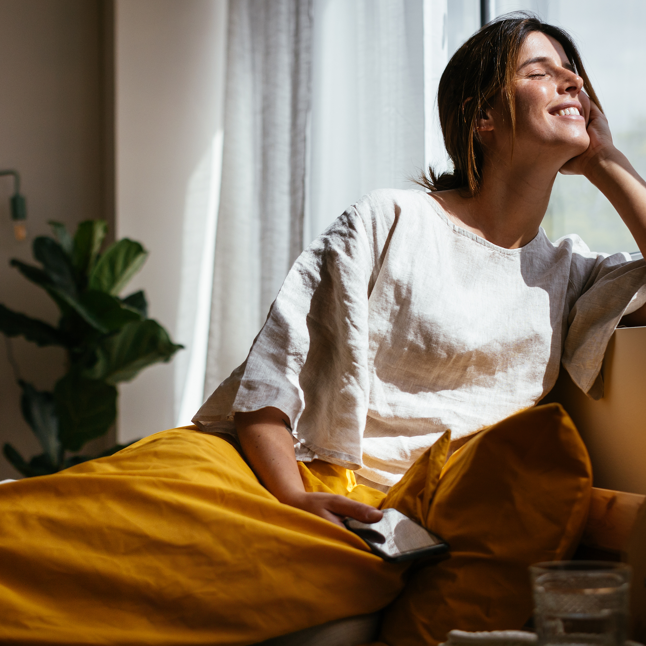 Woman sitting on a couch with yellow cushions, holding a book, in a bright room with natural light.