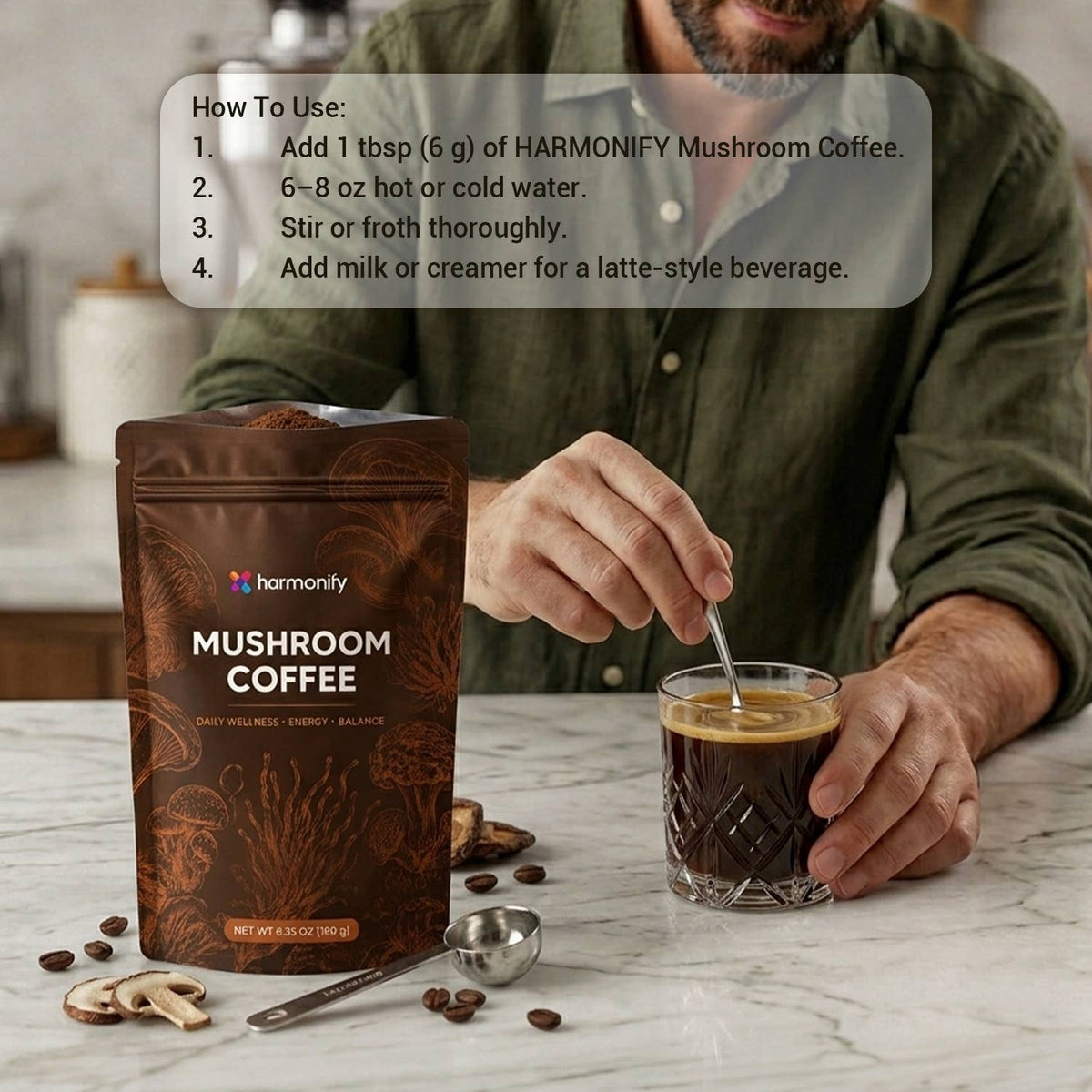 A man in a green shirt stands at a kitchen counter holding a glass of coffee while using a spoon to stir a pouch of Harmony Mushroom Coffee. The brown stand-up pouch is labeled 'Harmony Mushroom Coffee' and is shown on a marble countertop with coffee beans and a metal scoop nearby.