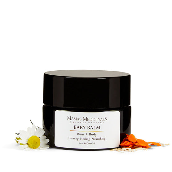 A jar of Mama's Medicinals Baby Bum + Body Balm with a white label, surrounded by a chamomile flower and some petals on a white background.