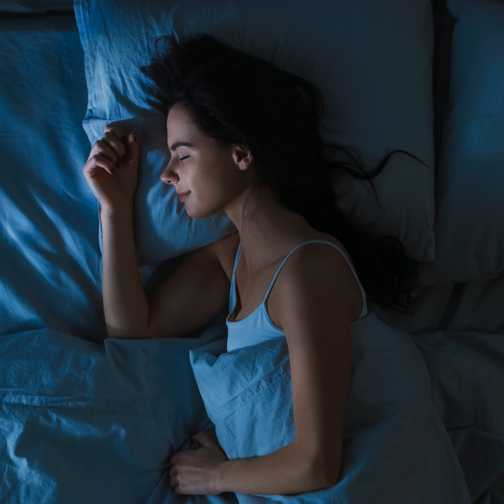 Woman lying in bed under blue sheets and pillowcases in a dimly lit room.