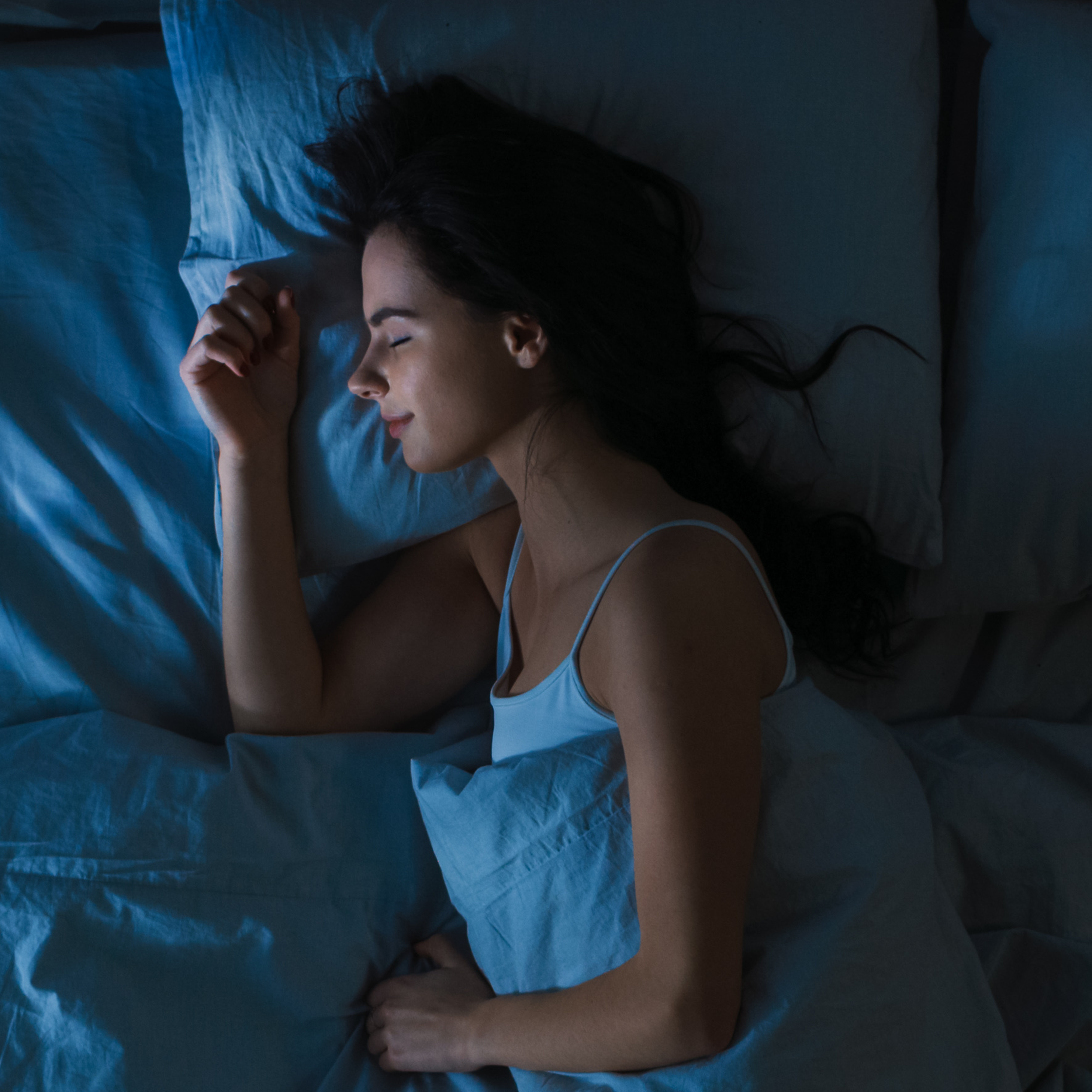 Woman lying in bed under blue sheets and pillowcases in a dimly lit room.