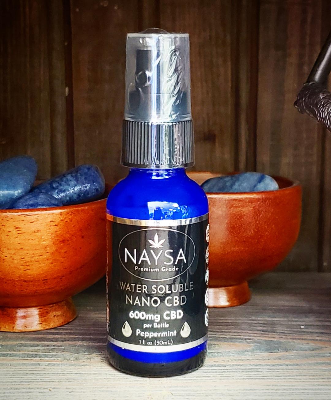 A blue bottle of Naysa Water Soluble CBD oil with a spray nozzle, displayed on a wooden surface with decorative items in the background.