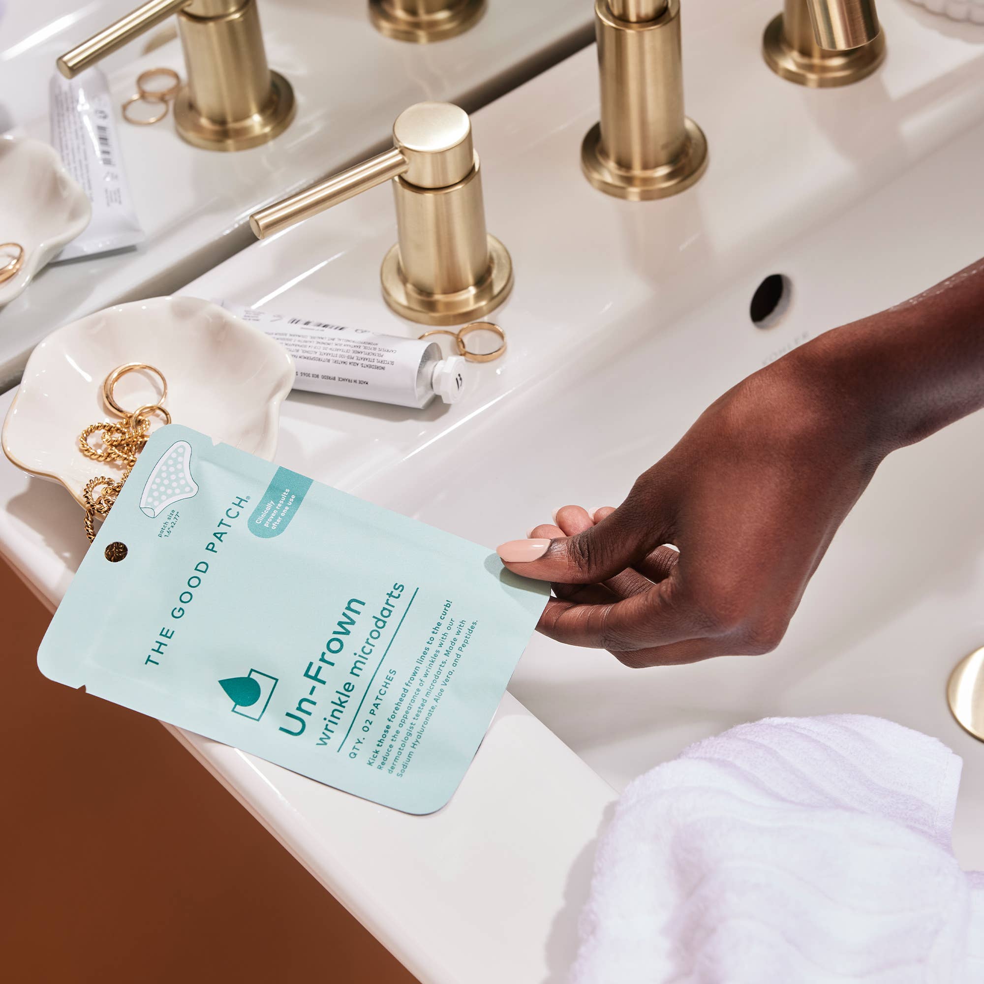 Hand holding a skincare product labeled 'The Good Patch' over a bathroom sink.