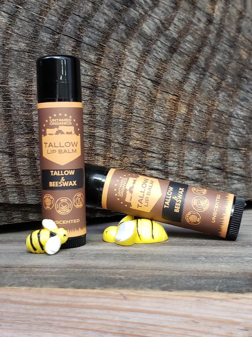Two sticks of lip balm with brown packaging, labeled 'TALLOW BEESWAX', one standing upright and the other lying down, with a small yellow object, possibly a honey dipper, in front of them, all placed on a wooden surface.